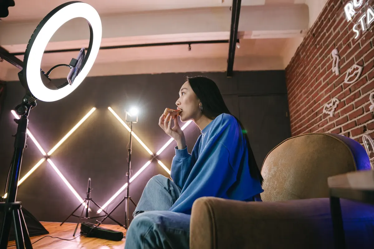 Asian woman content creator eating pizza while recording a video indoors with ring light.