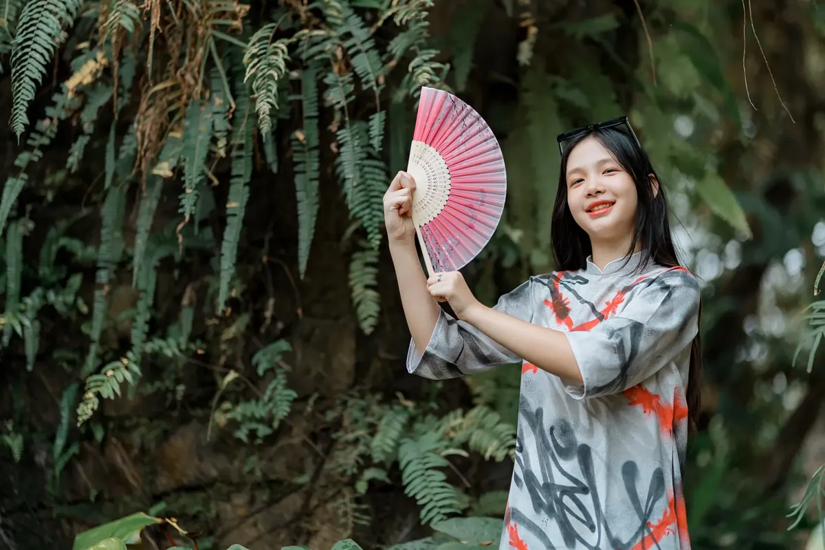 A young woman holding a fan smiles in a lush green forest, embodying joy and tranquility.
