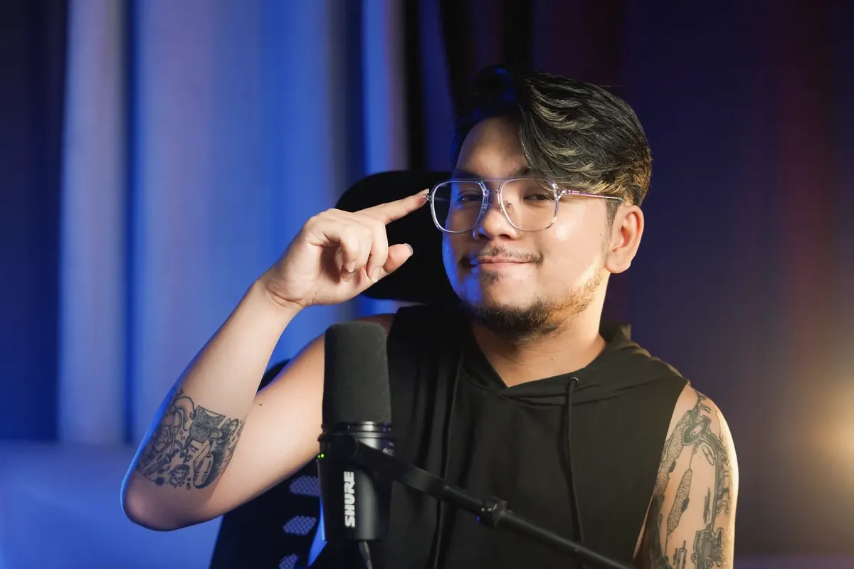 Young man with tattoos and eyeglasses sitting indoors, speaking into a microphone.