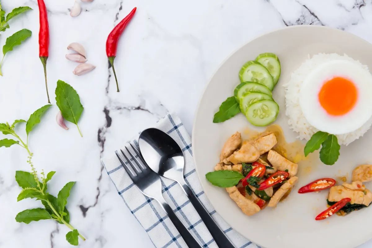 Close-up of a Thai meal with rice, chicken, and vegetables on a marble background.
