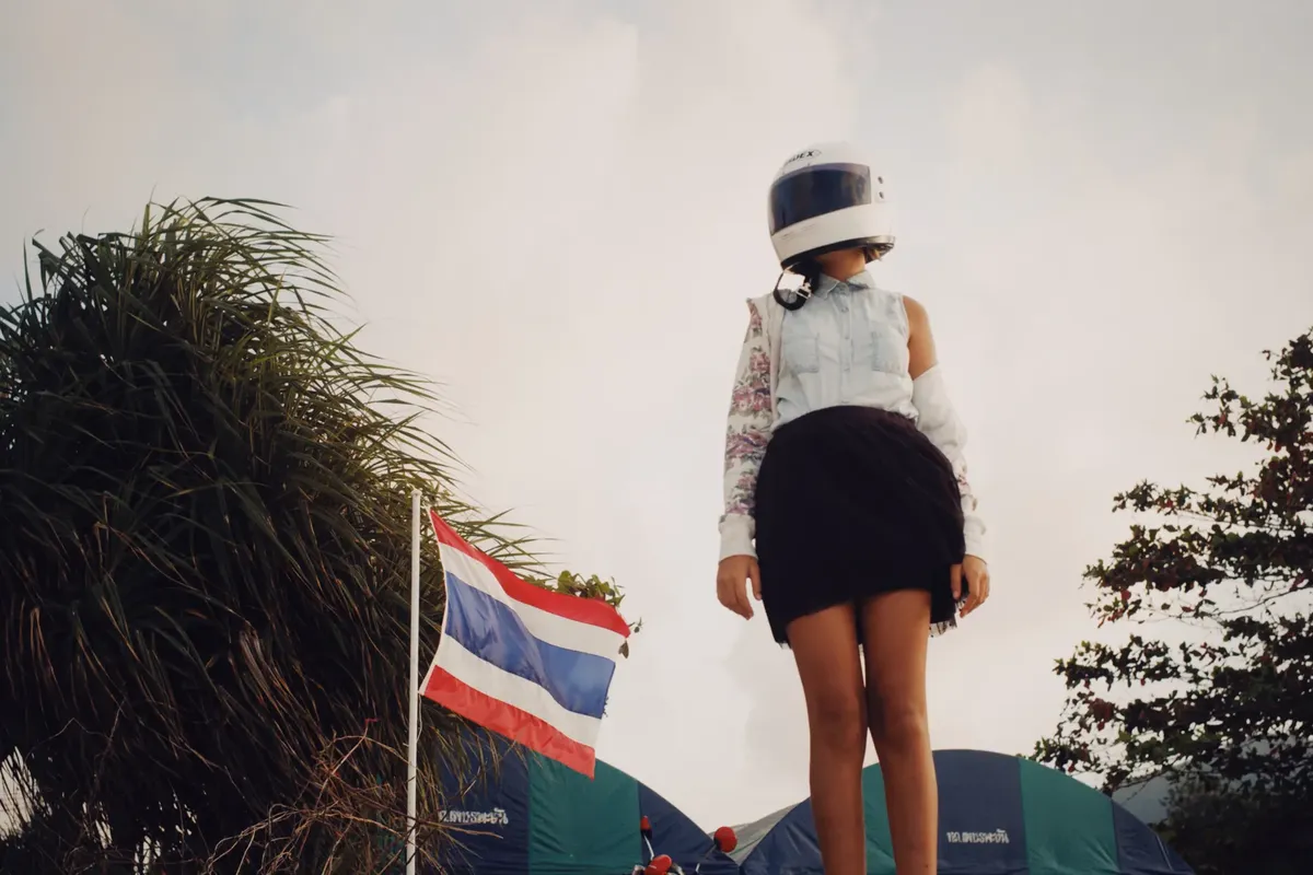 A woman stands outdoors in a helmet beside a waving Thai flag, captured from a low-angle perspective.